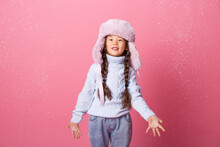 Cute Little Asian Girl In Winter Clothes Blows Snow From Palms. Winter End, Pink Background, Space For Text