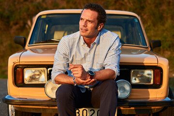 Sticker - Closeup shot of a man sitting in front of his old vintage car. Serious look on a curly model face