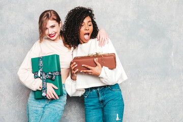 Two young beautiful smiling international hipster female in trendy clothes.Sexy carefree women posing near gray wall.Positive models hugging and giving each other gift boxes.Christmas, x-mas, concept