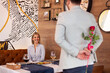 Beautiful girl is sitting at the table in restaurant and smiling while a man is holding rose behind his back. focus on woman.