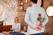 Beautiful girl is sitting at the table in restaurant and smiling while a man is holding rose behind his back. focus on flower.