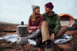 © StratfordProductions - Closeup of kettle on gas stove during camping with smiling couple looking at each other during winter - young adventurous couple camping and making coffee