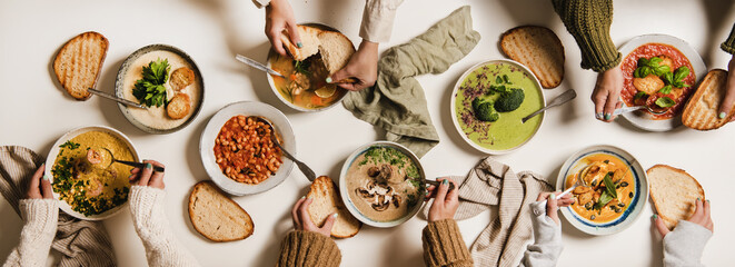 People eating Autumn and Winter creamy vegan soups, fall and winter vegetarian food menu. Flat-lay of peoples hands eating homemade soup with fresh bread over white table background, top view