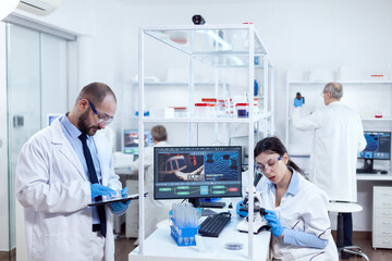 Scientist analysing test results on tablet pc. Team of researchers doing pharmacology engineering in sterile laboratory for healthcare industry with african assistant in the background.