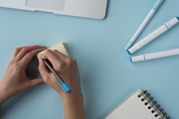 Poster - High angle shot of a person writing on sticky notes and a journal, pens, and laptop on the table