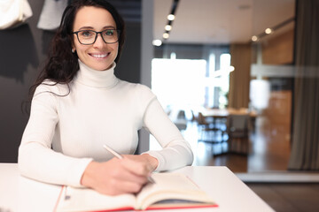 Brunette woman with glasses smiling and holding ballpoint pen over notebook