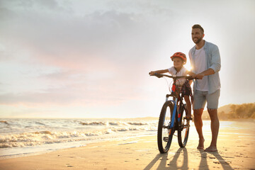 Wall Mural - Happy father teaching son to ride bicycle on sandy beach near sea at sunset