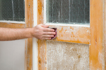 Young adult man hand using sandpaper and sanding wooden door from old white color. Closeup. Renovation process.