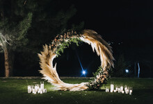 A Round, Luminous Wedding Arch, Decorated With Reeds And Glass Candlesticks With Burning Candles And Lamps, Stands On Green Grass, Against The Backdrop Of Nature In The Forest At Night.