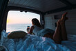 © Alexandra C. Ribeiro/Westend61 - Woman taking photo of sunset through phone while lying in camper van at beach