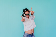 Happy Fashionable Woman Showing Peace Sign While Listening Music Through Headphones Against Turquoise Background