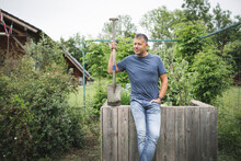 Handsome Man Holding Spade Looking Away While Leaning On Wooden Raised Bed At Vegetable Garden