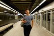 © VALENTINA BARRETO STUDIO/Westend61 - Handsome young man holding smart phone while walking with hand in pocket at illuminated subway platform