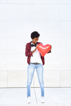 Happy Afro Woman Looking At Red Heart Shape Balloon Against White Wall