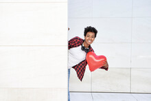 Happy Young Woman Holding Red Heart Shape Balloon While Standing Behind White Wall