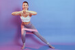 Winsome sporty beautiful dark haired woman with hair in ponytail stretching her body and doing side lunge exercises with fitness rubber band isolated over color background.