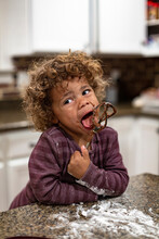 Cute Mixed-race Boy Licking The Blender Beater From A Bowl Of Brownie Cake Batter As He Makes A Mess In The Kitchen. Child Having Fun At Home Baking And Cooking