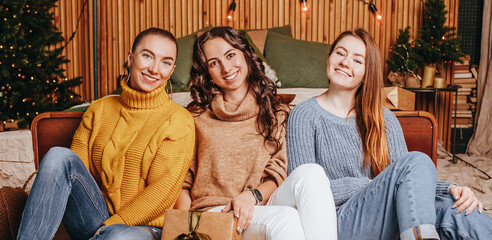 Wall Mural - Three beautiful cheerful happy young girls girlfriend give Christmas gifts on the background of a new year tree at home