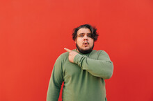 Surprised Corpulent Hispanic Guy Wearing A Green Sweatshirt Is Pointing His Finger Backwards On A Red Background, Looking Shocked And Astonished.