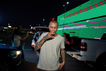 A young dark-skinned casualy dressed tattooed man smoking a cigarette with his friends cars and a led screen on a background