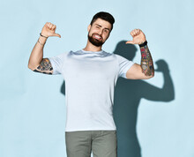 Brutal Bearded Man Fun Guy With Tattoo On Arms In White Shiny Silk T-shirt Looking At Camera Pointing Thumbs At Himself And Winks Over White Background. Hipster Stylish Look For Free Lifestyle Concept