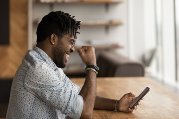 Wall Mural - Overjoyed young biracial man look at cellphone screen triumph win lottery online. Happy excited African American male feel euphoric with good amazing unbelievable news or message on smartphone.