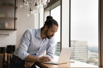 Focused millennial African American businessman look at laptop screen consult client or customer online. Serious young biracial man work on computer in office, browse wireless internet on gadget.