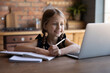 Smiling little girl watching online lesson, webinar, internet course, writing notes, homeschooling concept, happy child sitting at table in kitchen at home, looking at computer screen, studying