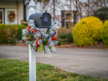 View Of US Mailbox Decorated With  Colorful Christmas Wreath; House And Bushes In Background