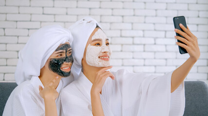 Two happy Asian girls in white bathrobes with facial mask taking photo on smartphone together in living room.