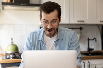 Wall Mural - Close up amazed young man wearing glasses using laptop in kitchen, surprised businessman or student with open mouth looking at laptop screen, reading good unexpected news, promotion or lottery win