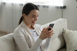 © fizkes - Laughing young woman looking at phone screen, sitting on couch, happy smiling girl having fun with smartphone, watching funny video in social network, reading news or message, enjoying leisure time