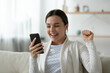 © fizkes - Close up overjoyed young woman looking at phone screen, showing yes gesture, reading good news in message, celebrating success, great shopping offer, lottery win, sitting on couch at home