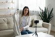 © fizkes - Young woman vlogger shooting clothing review video, using smartphone on tripod, speaking, attractive girl influencer blogger holding demonstrating clothes, sitting on cozy couch in living room