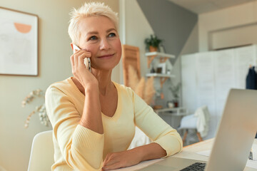 Beautiful female freelancer calling client using mobile working on portable computer. Successful woman entrepreneur on retirement having phone conversation indoors, using laptop for distant work