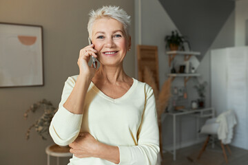 Modern beautiful retired woman posing in stylish living room with smart phone at her ear, smiling at camera, having conversation with friend, listening to news or gossip. Female pensioner calling son