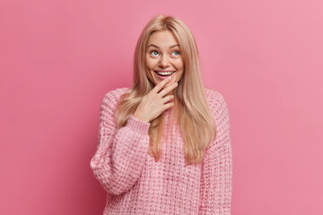 Wall Mural - Joyful blonde woman with thoughtful happy expression concentrated above smiles broadly shows white teeth wears warm winter sweater isolated over pink background. Positive human emotions concept.