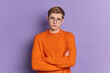 © WHstudio Leushin N - Displeased handsome young man has pity expression looks dissatisfied at camera keeps arms folded wears round spectacles and orange jumper isolated over purple background. Face expressions concept