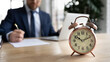© fizkes - Close up alarm clock on desk, businessman wearing suit using laptop, working with correspondence on background, entrepreneur involved in working process, workday, time management concept