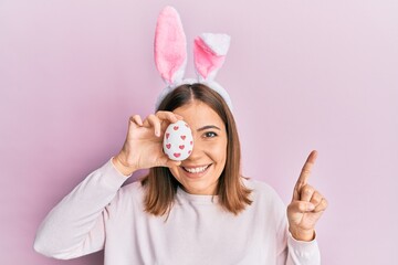 Sticker - Young beautiful woman wearing cute easter bunny ears holding egg smiling with an idea or question pointing finger with happy face, number one