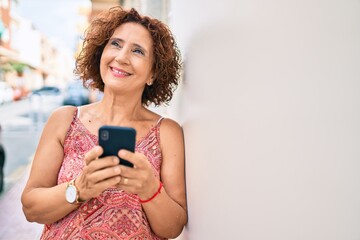 Wall Mural - Middle age woman using smartphone leaning on the wall at street of city.