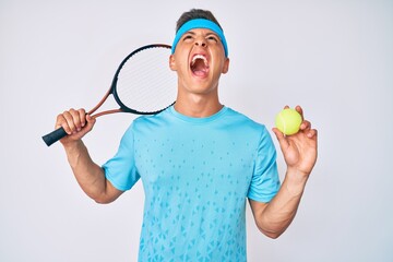 Poster - Young hispanic boy playing tennis holding racket and ball angry and mad screaming frustrated and furious, shouting with anger looking up.