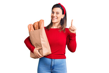 Wall Mural - Young beautiful woman holding paper bag with bread surprised with an idea or question pointing finger with happy face, number one