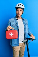 Sticker - Young man standing on scooter wearing helmet holding first aid kit in shock face, looking skeptical and sarcastic, surprised with open mouth