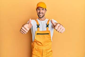 Poster - Young handsome man wearing handyman uniform over yellow background approving doing positive gesture with hand, thumbs up smiling and happy for success. winner gesture.