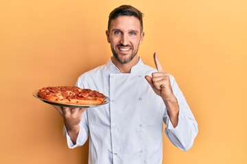 Wall Mural - Handsome man with beard professional cook holding italian pizza smiling with an idea or question pointing finger with happy face, number one