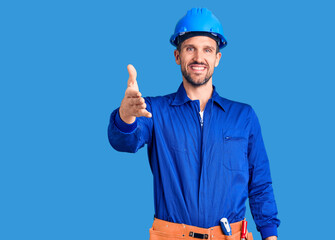 Canvas Print - Young handsome man wearing worker uniform and hardhat smiling friendly offering handshake as greeting and welcoming. successful business.