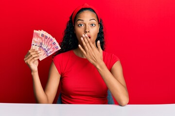 Canvas Print - Young african american girl holding south african 50 rand banknotes sitting on the table covering mouth with hand, shocked and afraid for mistake. surprised expression