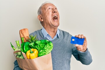 Canvas Print - Senior caucasian man holding groceries and credit card angry and mad screaming frustrated and furious, shouting with anger looking up.