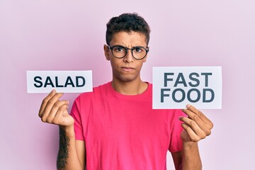 Sticker - Young handsome african american man holding salad and fast food message paper skeptic and nervous, frowning upset because of problem. negative person.